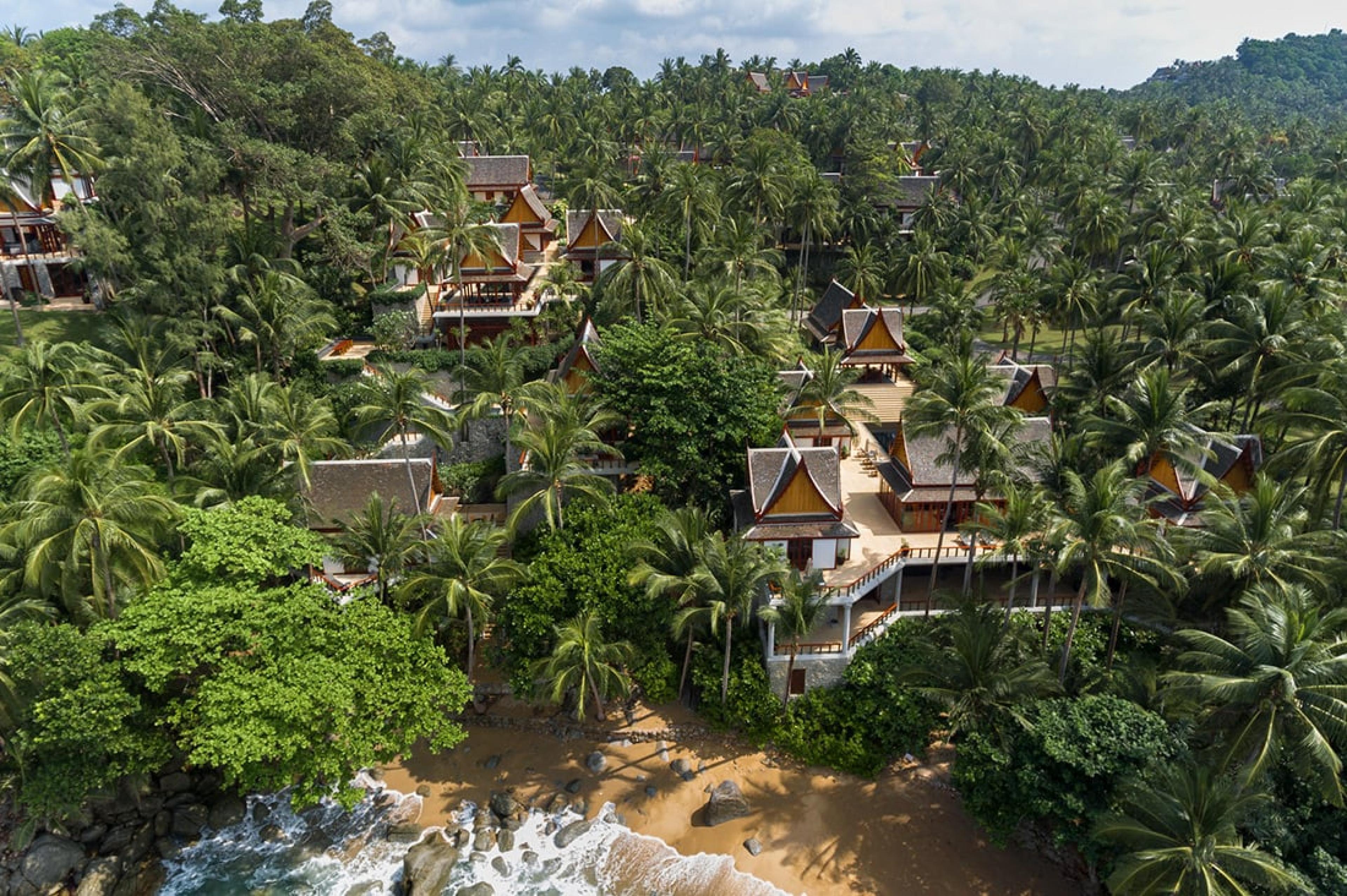 aerial view looking over beach towards tropical hill with thai-style huts comprising a luxury hotel