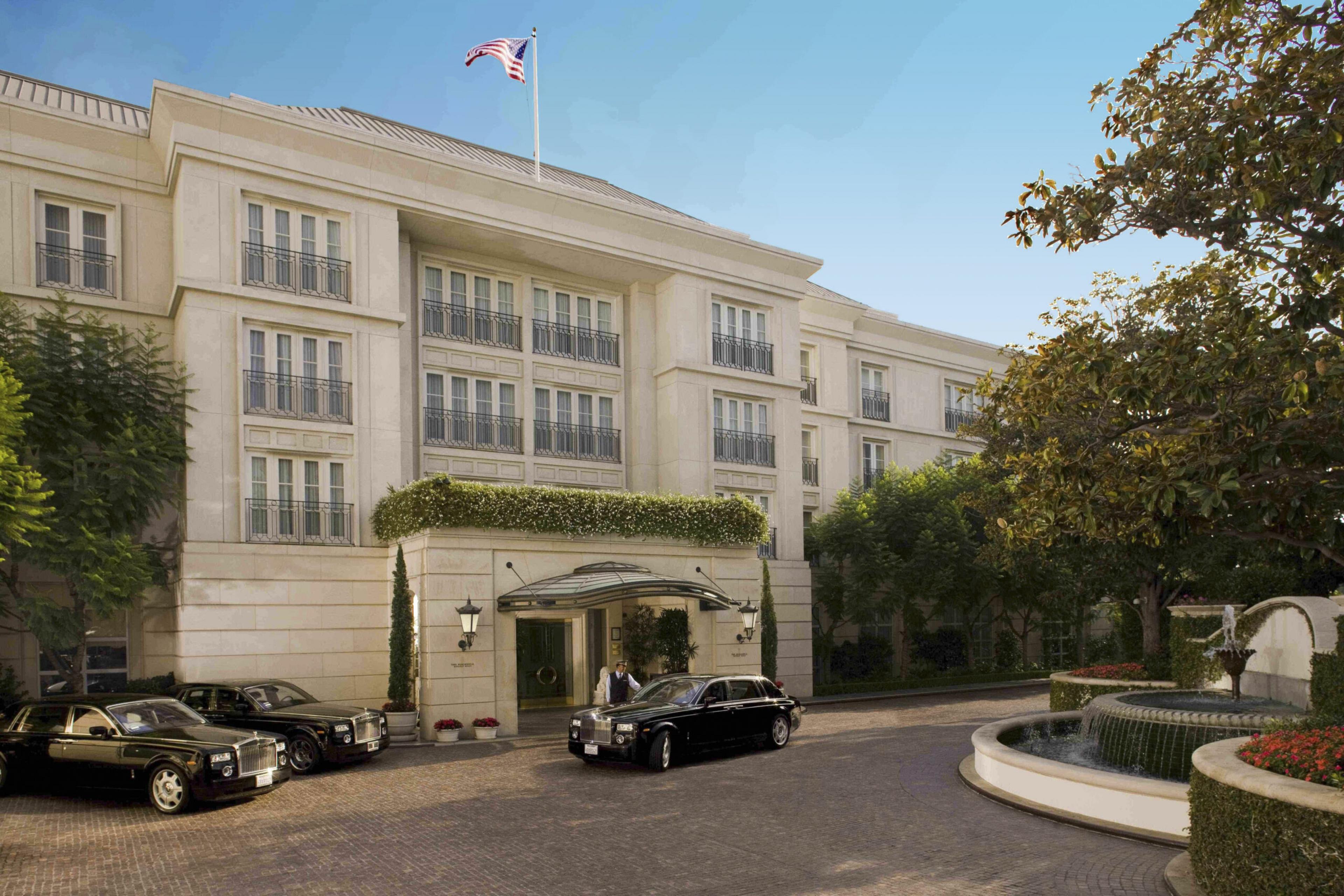 Front driveway and entrance of the hotel. The building is large and white and a worker opens a car door of a Rolls Royce