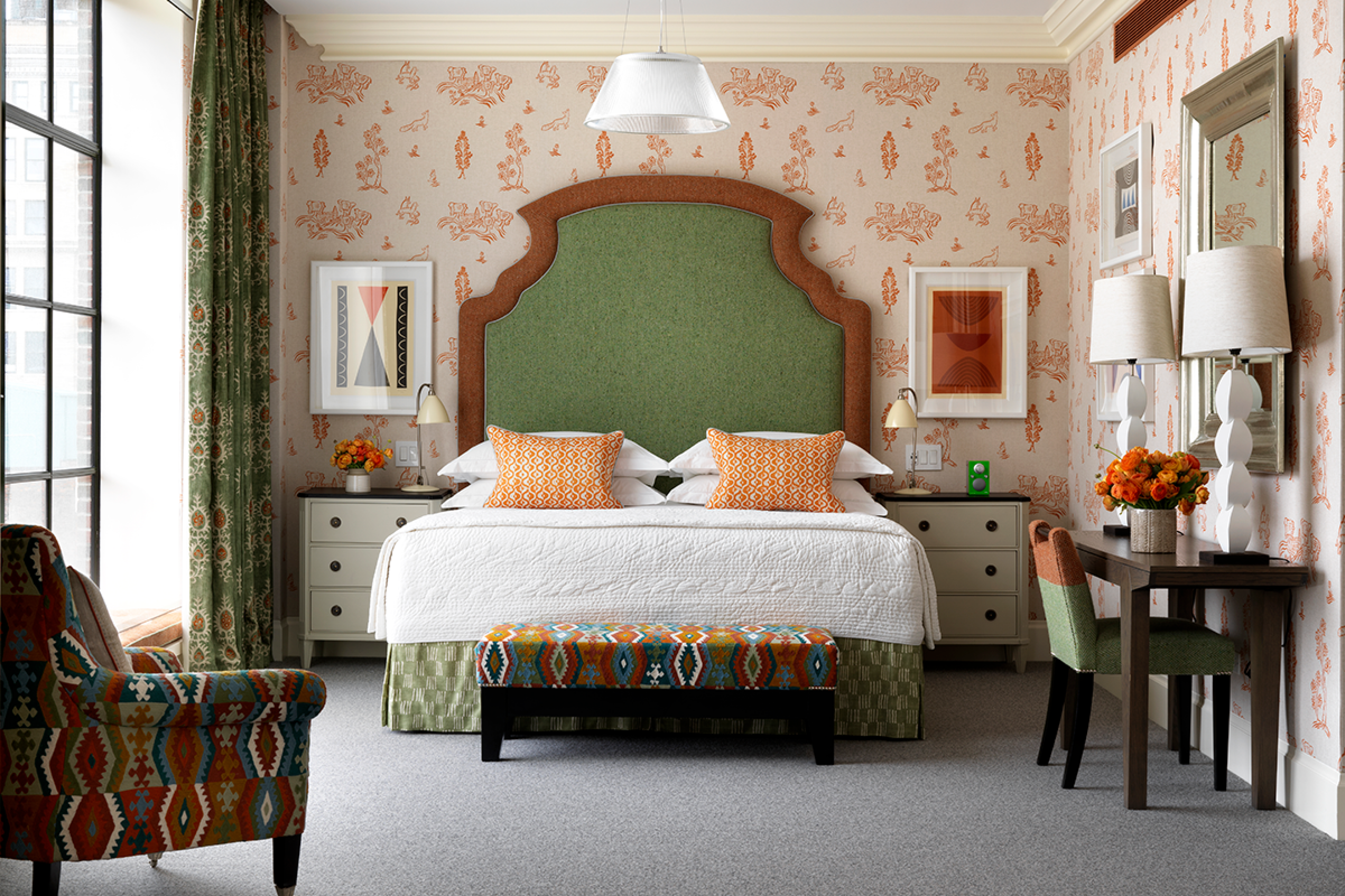 Colorful bedroom with orange wallpaper and pillows and large green headboard. Matching nightstands are on either side of bed and ottoman is at foot of bed