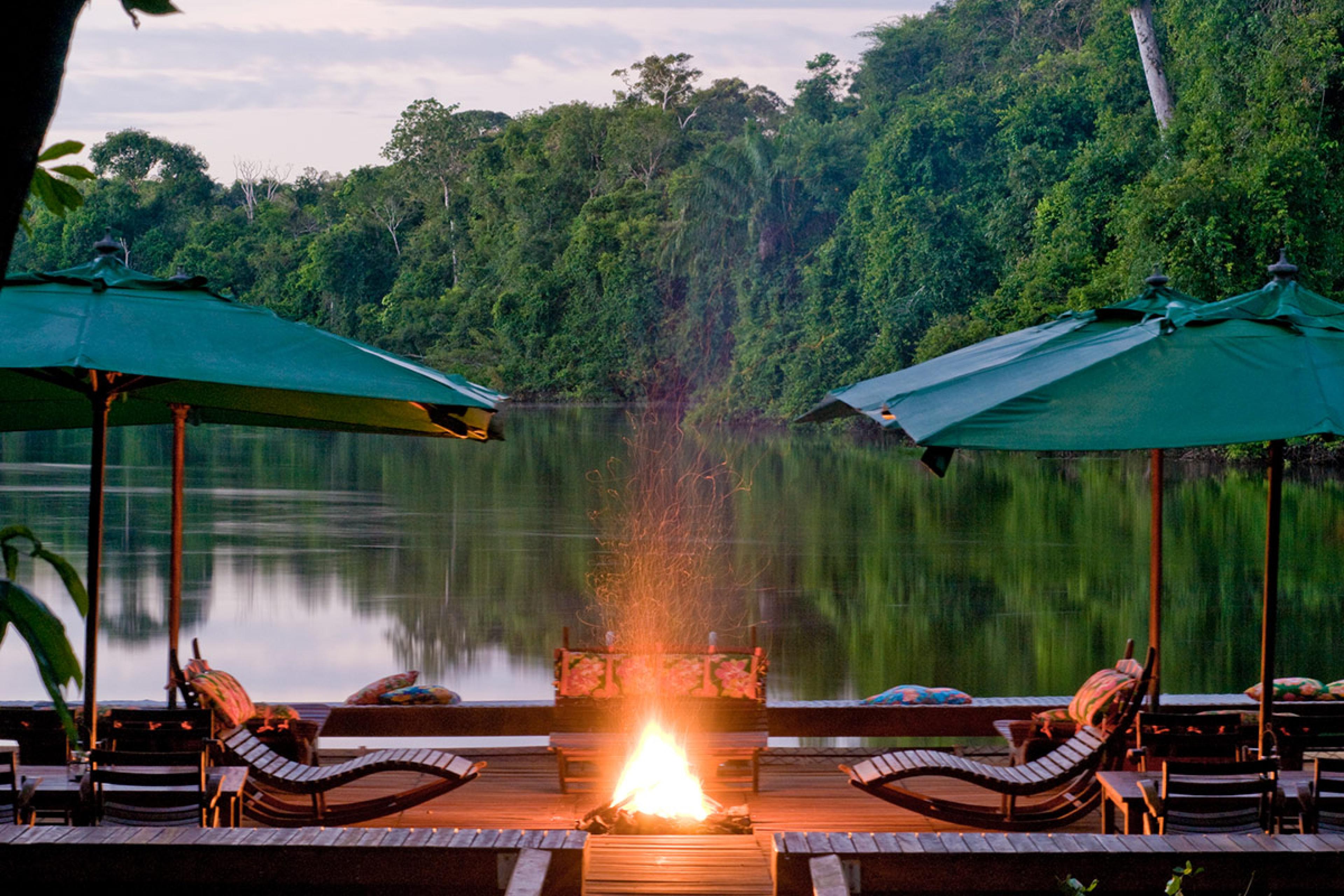 a fire pit on a floating dock with wooden lounge chairs and a green umbrellas at dusk along a river