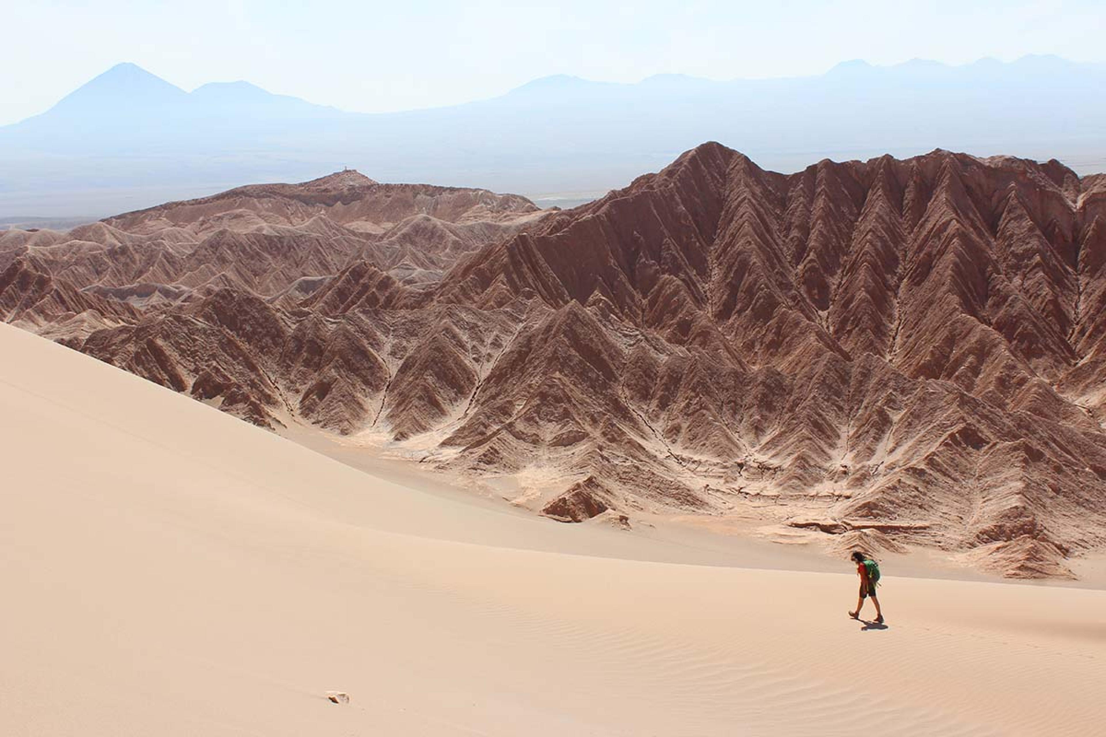 person walking in a desert with rippling mountains behind him