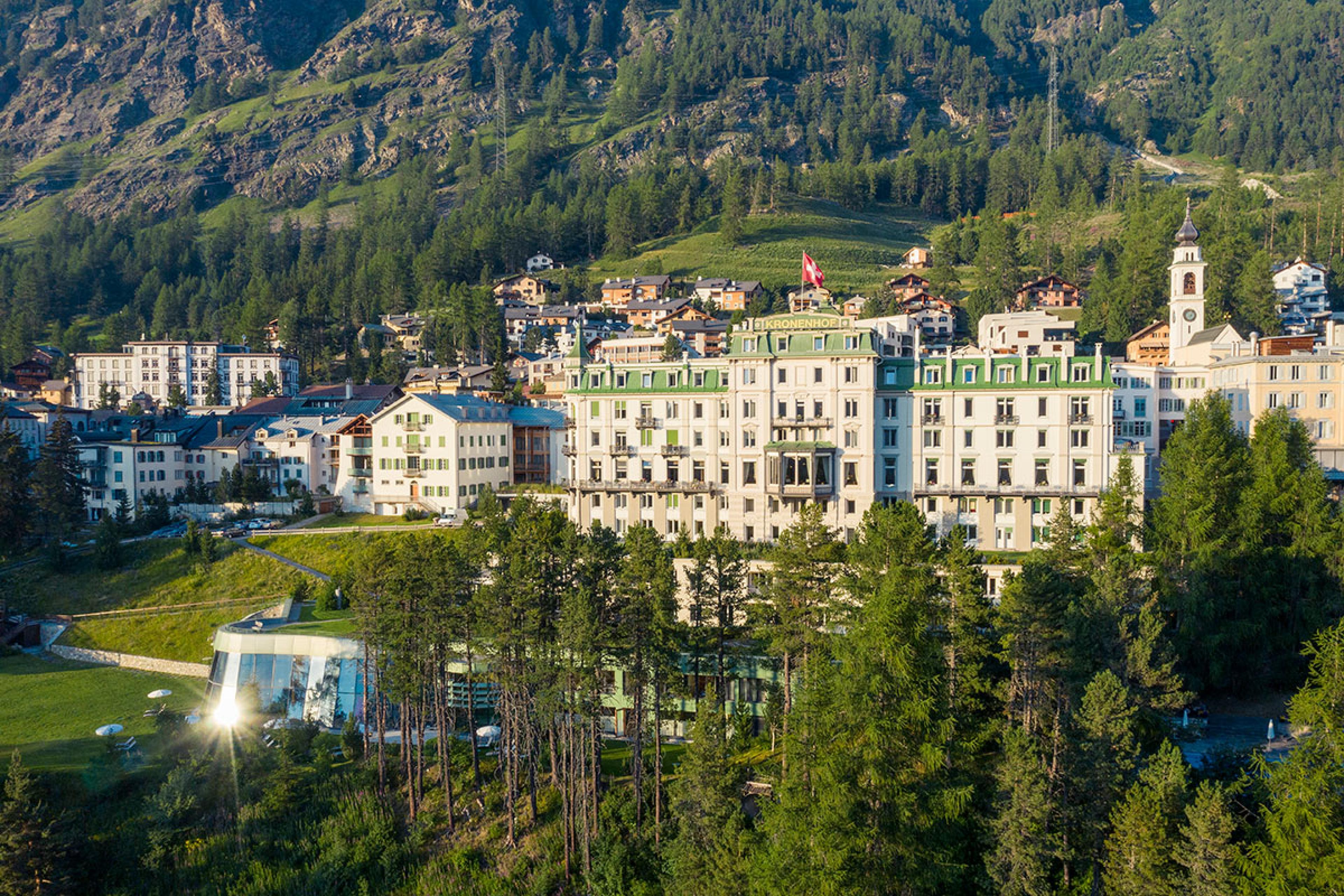 exterior of historic swiss hotel in summertime with green roof and white walls with mountain in background and trees in foreground