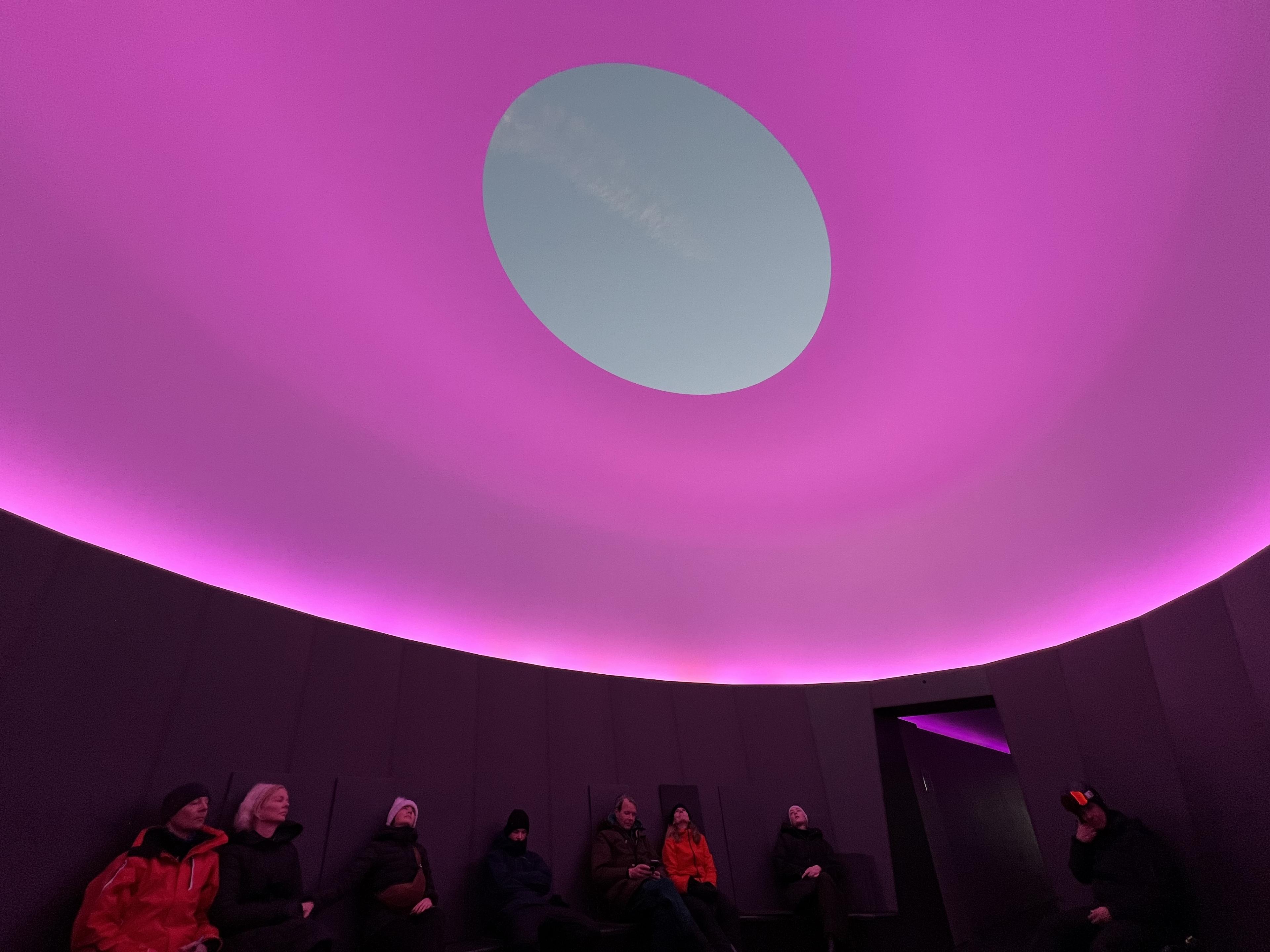 looking up at oval hole in the ceiling of james turrell skyspace during light show where ceiling and walls are lit in pink