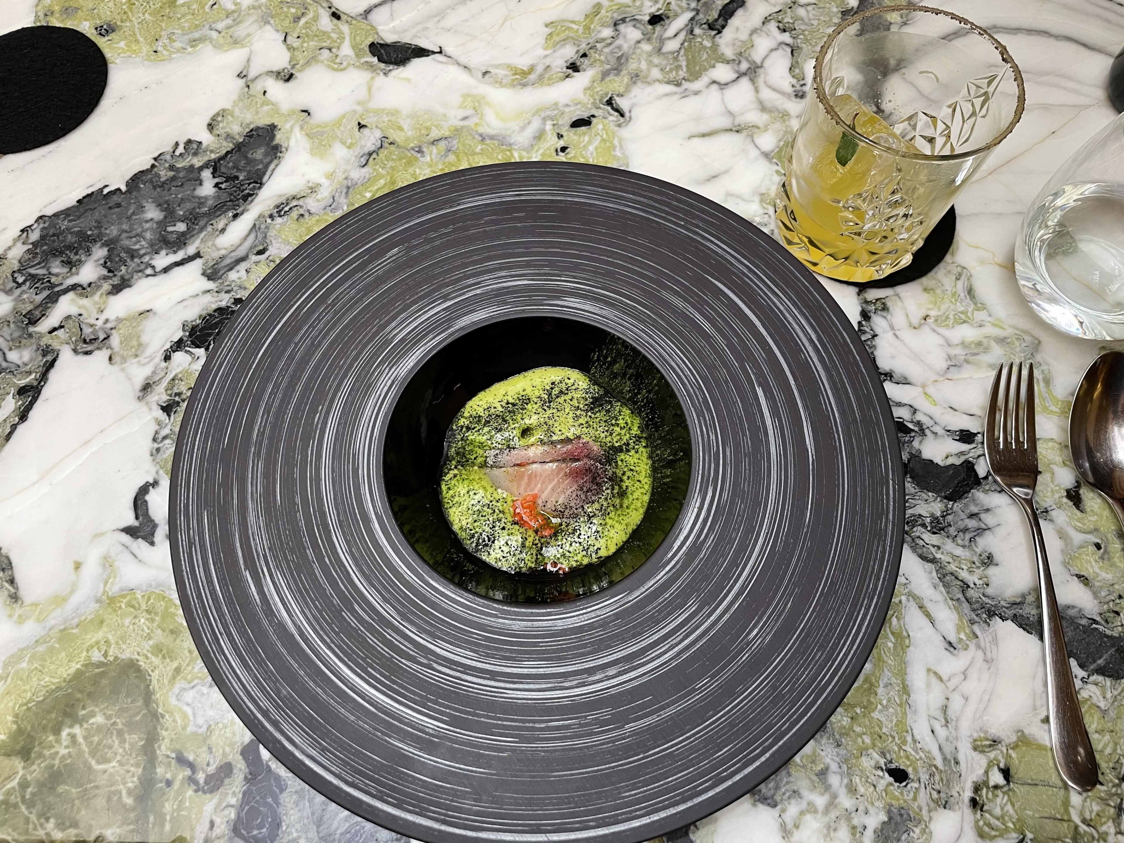 overview of circular gray plate on a stone table. inside plate is fish in a green sauce presented in a fine dining manner