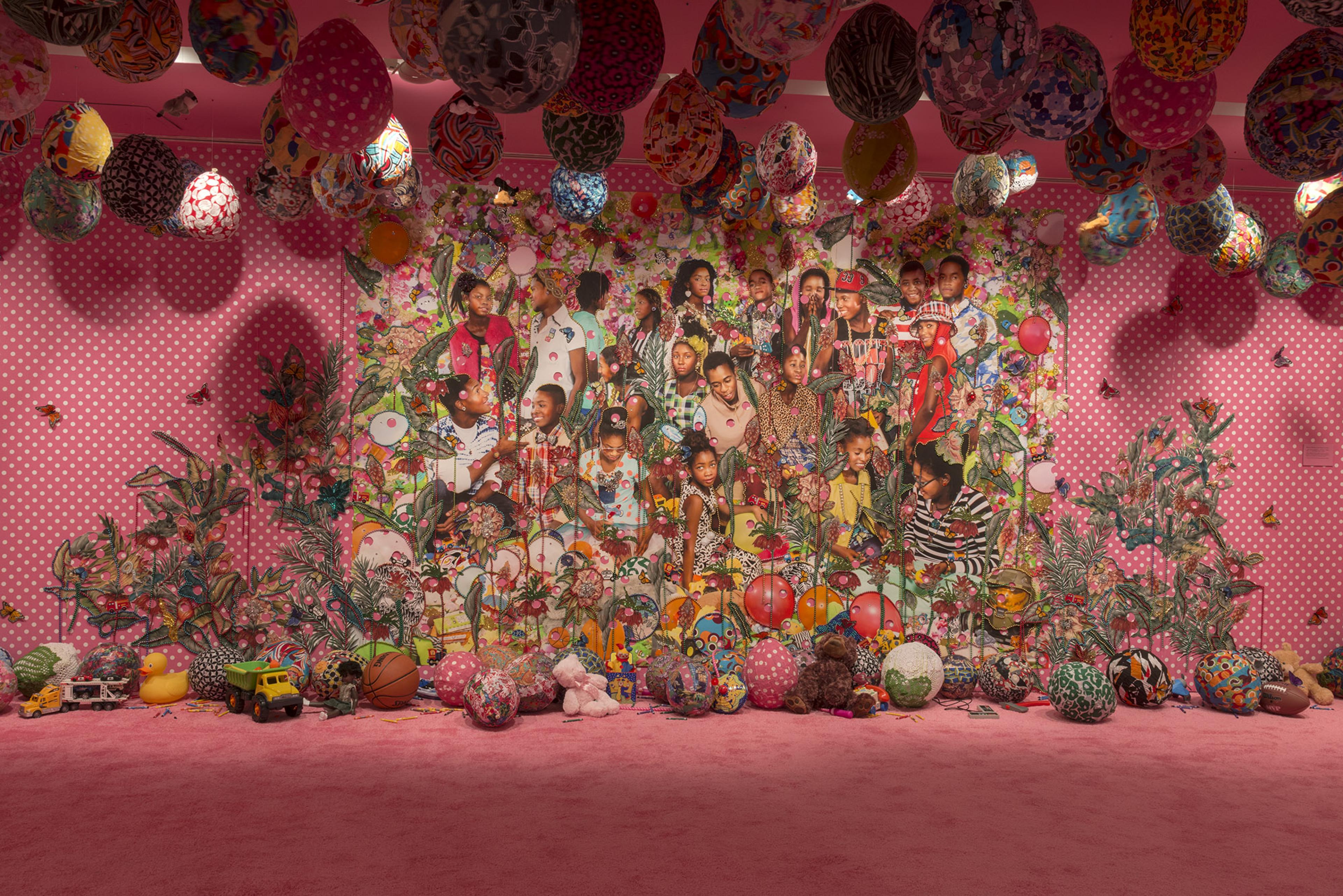 an art installation against a pink wall with a collage of photos of black children as well as mardi gras beads, balloons and teddy bears