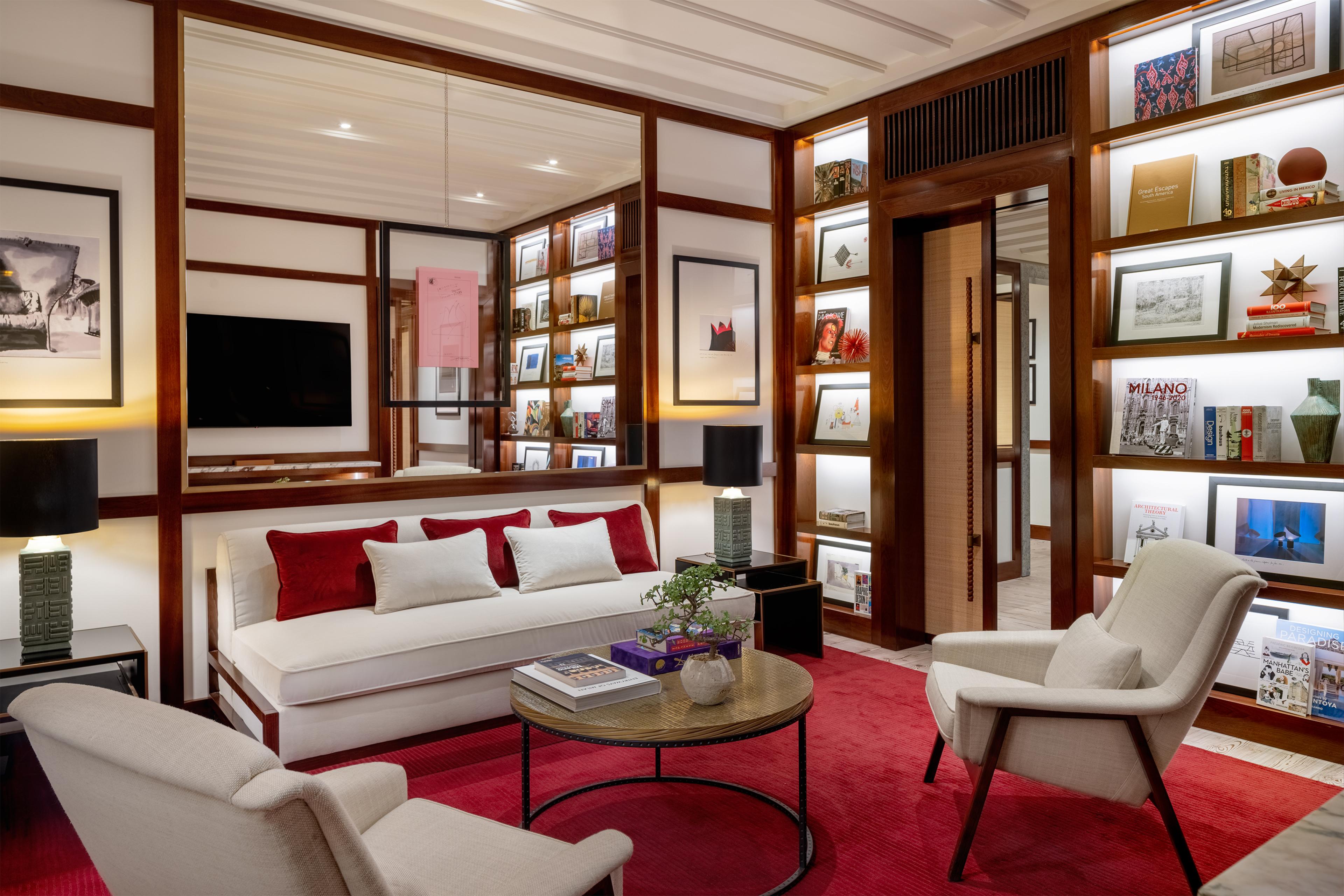 a hotel room living area with red accents, sleek white lighting and warm dark wood furnishings as well as well-lit bookcases 