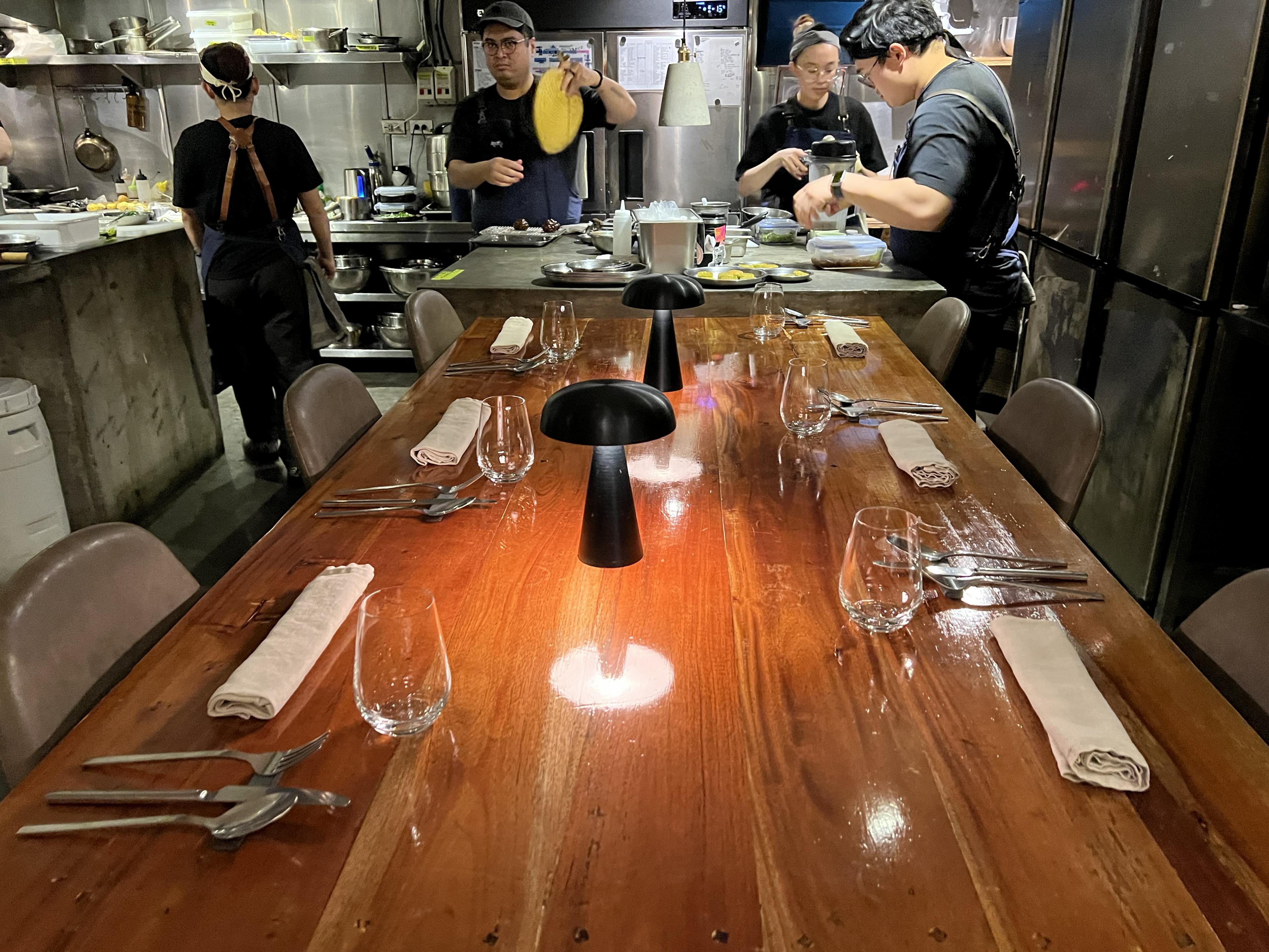 wooden table set for dinner inside restaurant kitchen where staff works at metal counters