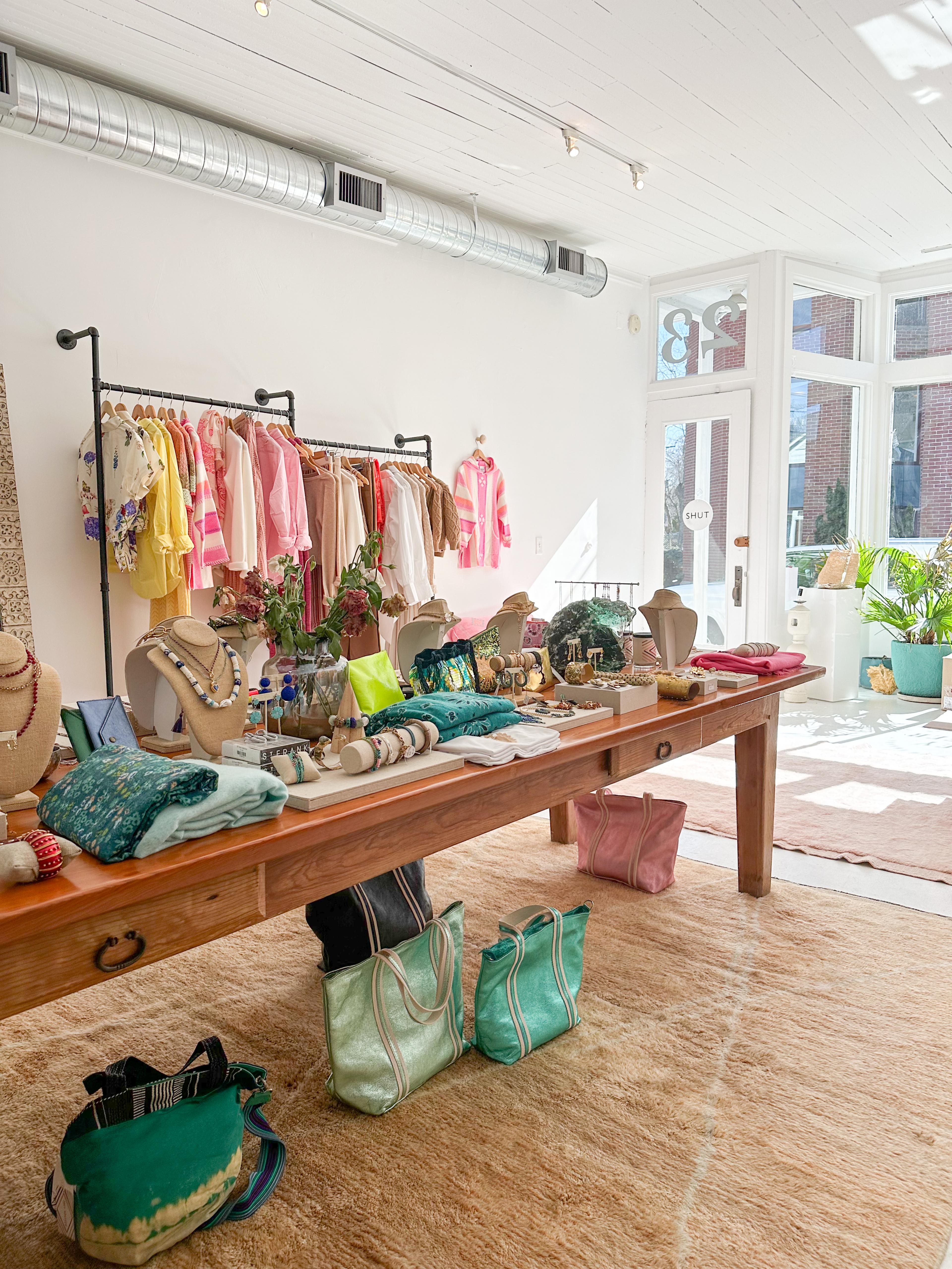 women's clothing store with pink and yellow clothes hanging from metal racks along white painted wall and also a wooden table in foreground with ladies' accessories