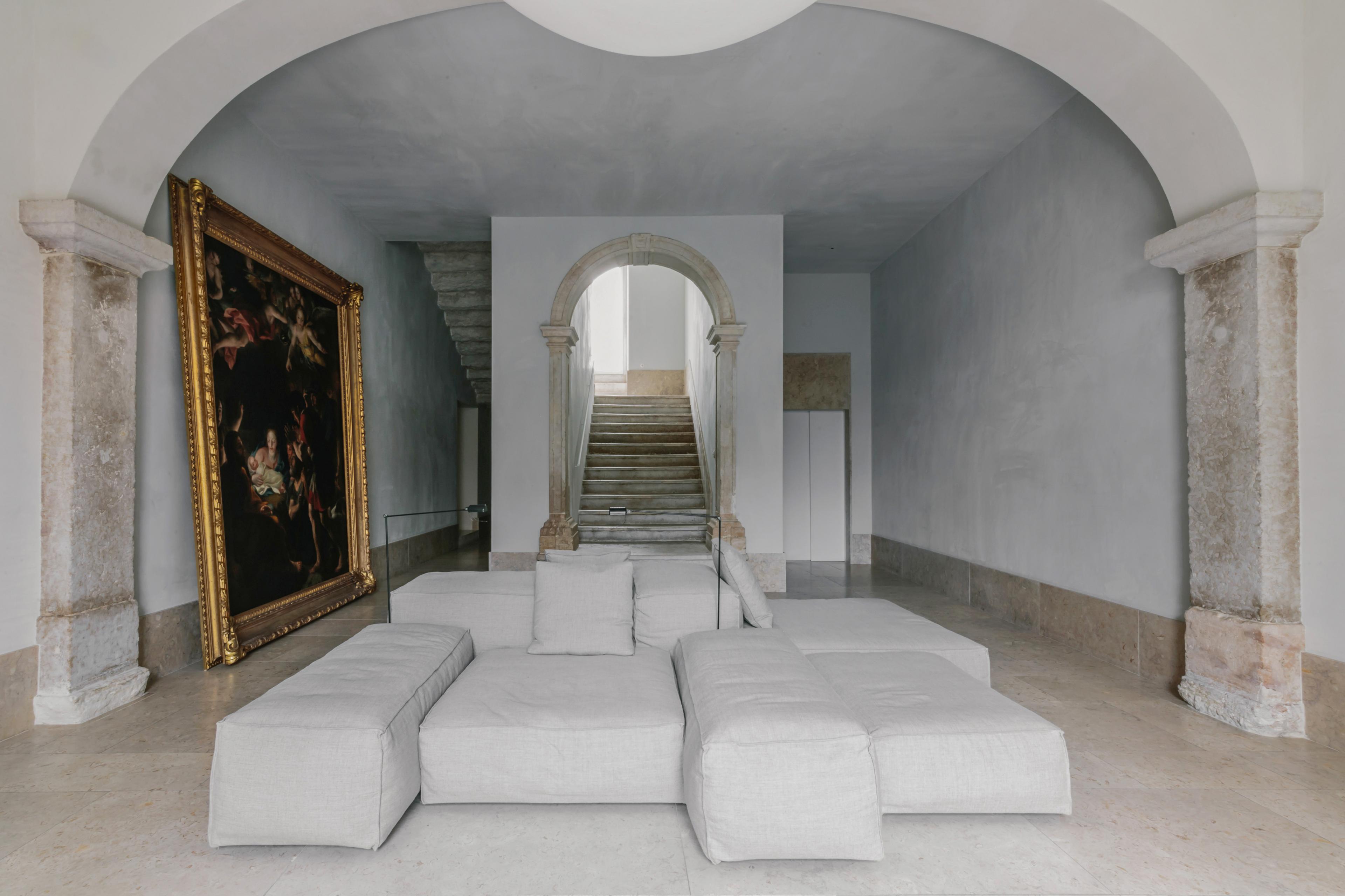 whitewashed walls and a vaulted ceiling in an old historic home in europe, with a cluster of white poofy-square-seats arranged in the center and a grand oil painting taking up the full wall on the left. a staircase is in the background