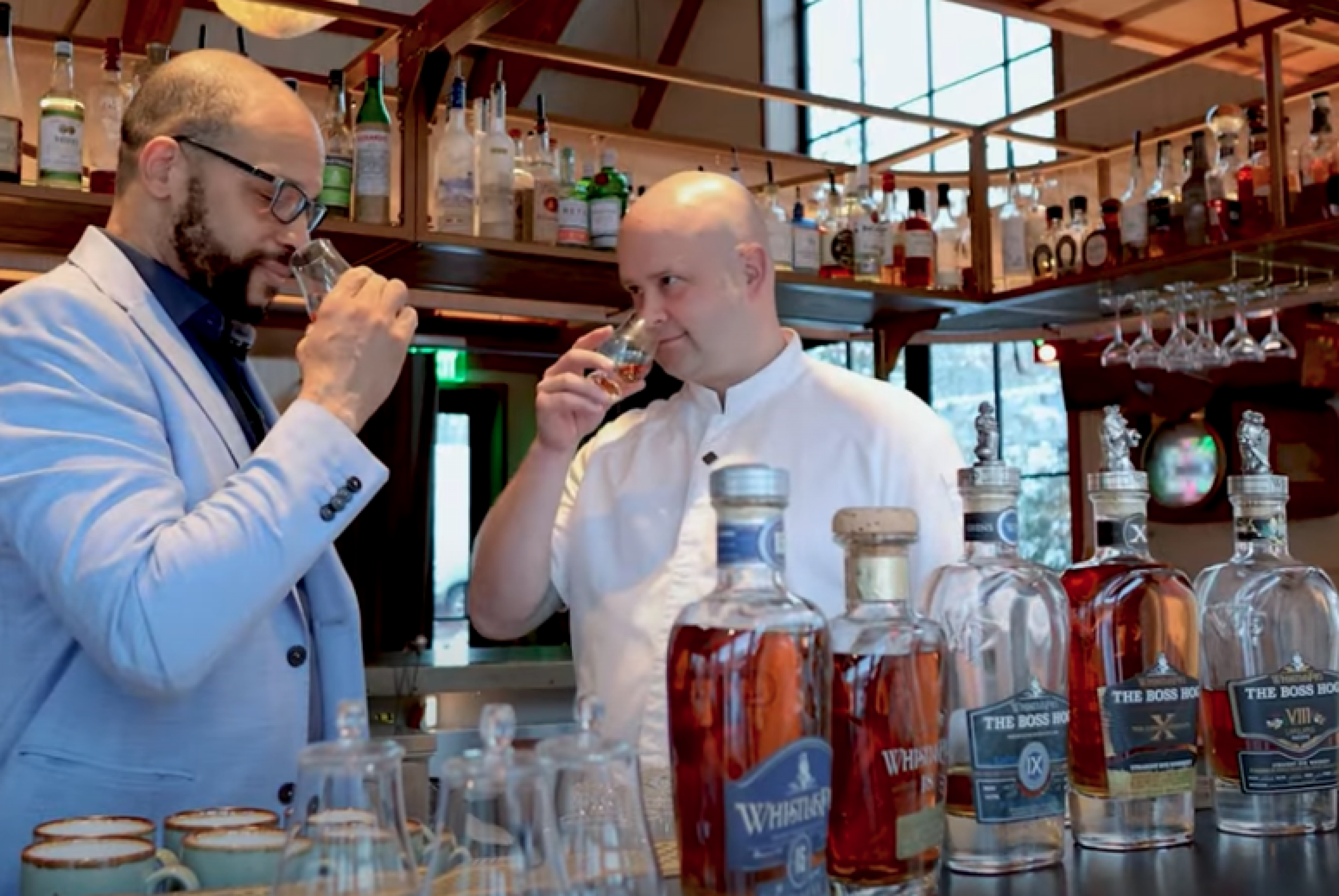 Two men smelling whiskey in snifters with bottles of whiskey in the background