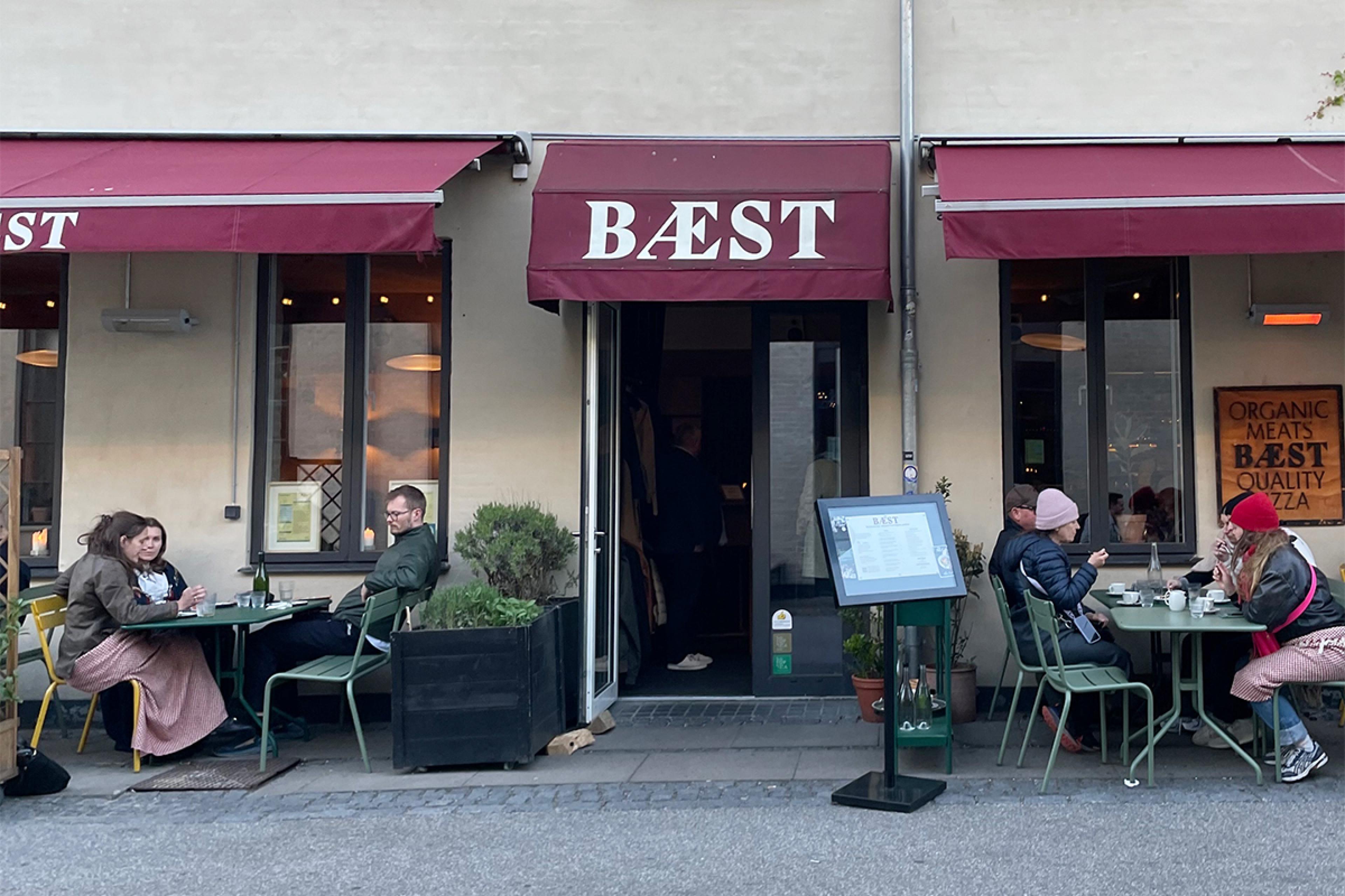exterior of restaurant in copenhagen with stucco walls and sign saying Baest above open door. People wearing clothing for a cold fall day are sitting at tables outside.