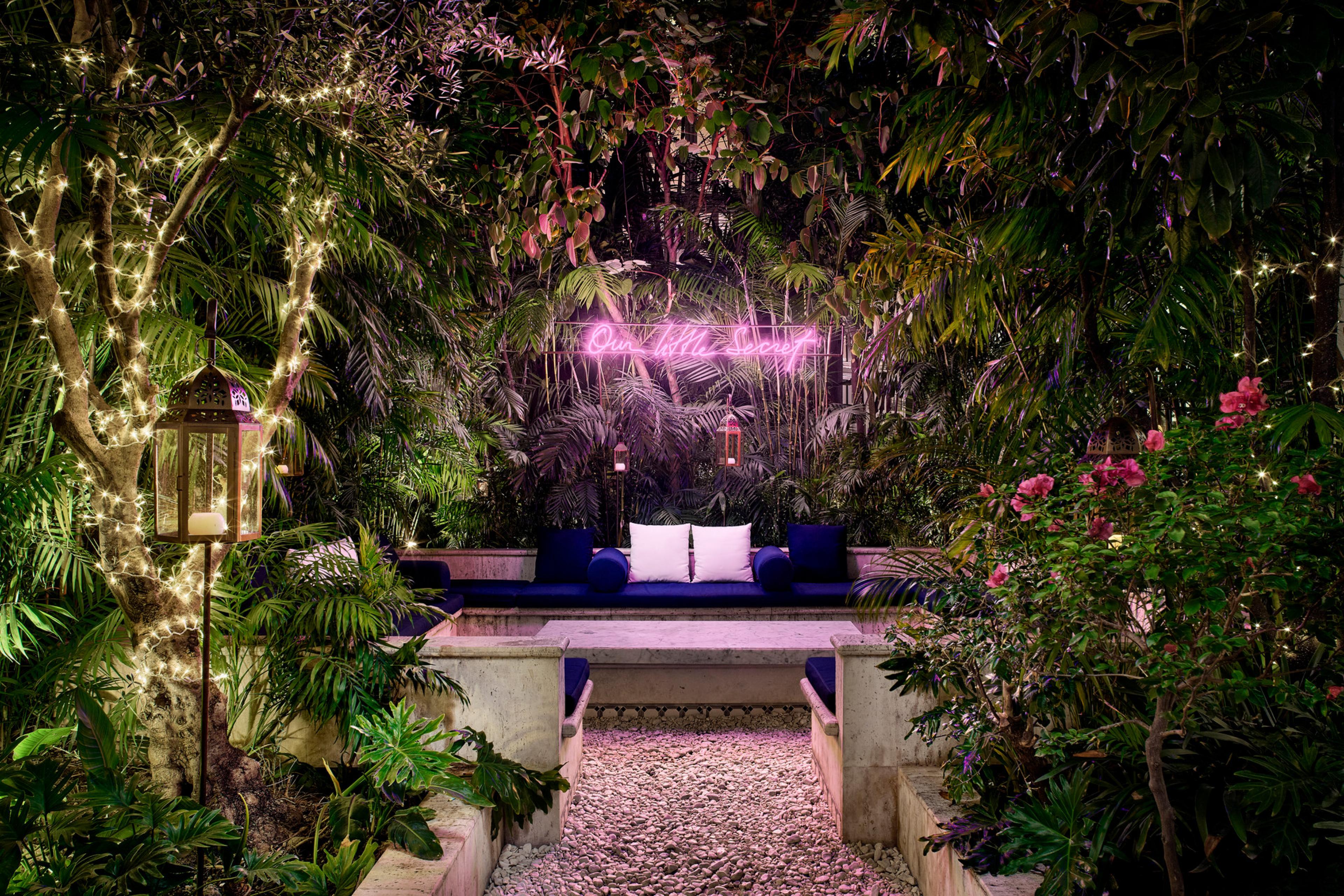 A small outdoor bench seat surrounded by lush greenery with a neon sign that reads "our little secret"