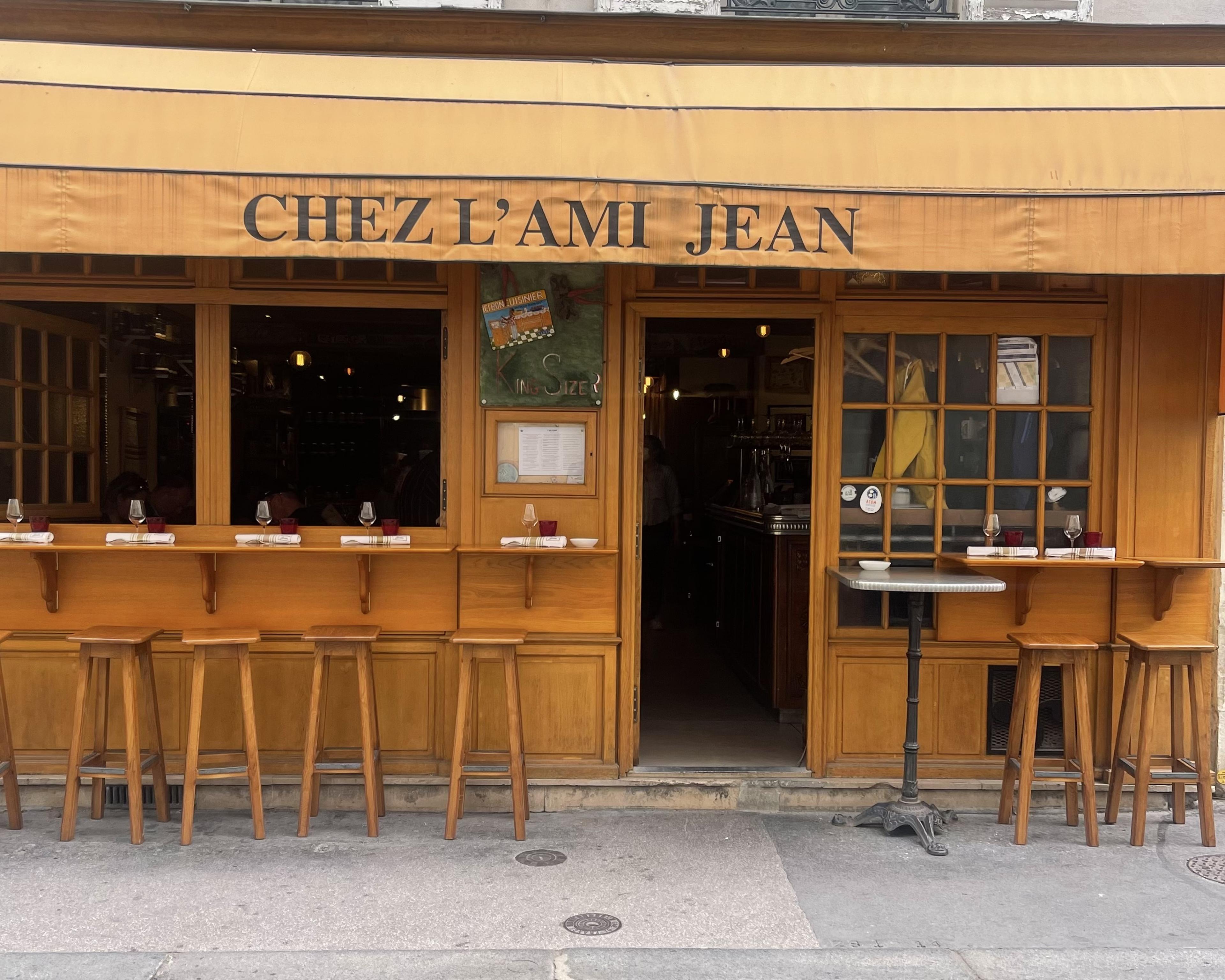 Restaurant with orange awning saying Chez L'Ami Jean with bar stools arranged in front of exterior windows so diners could eat at counters on exterior orange wall