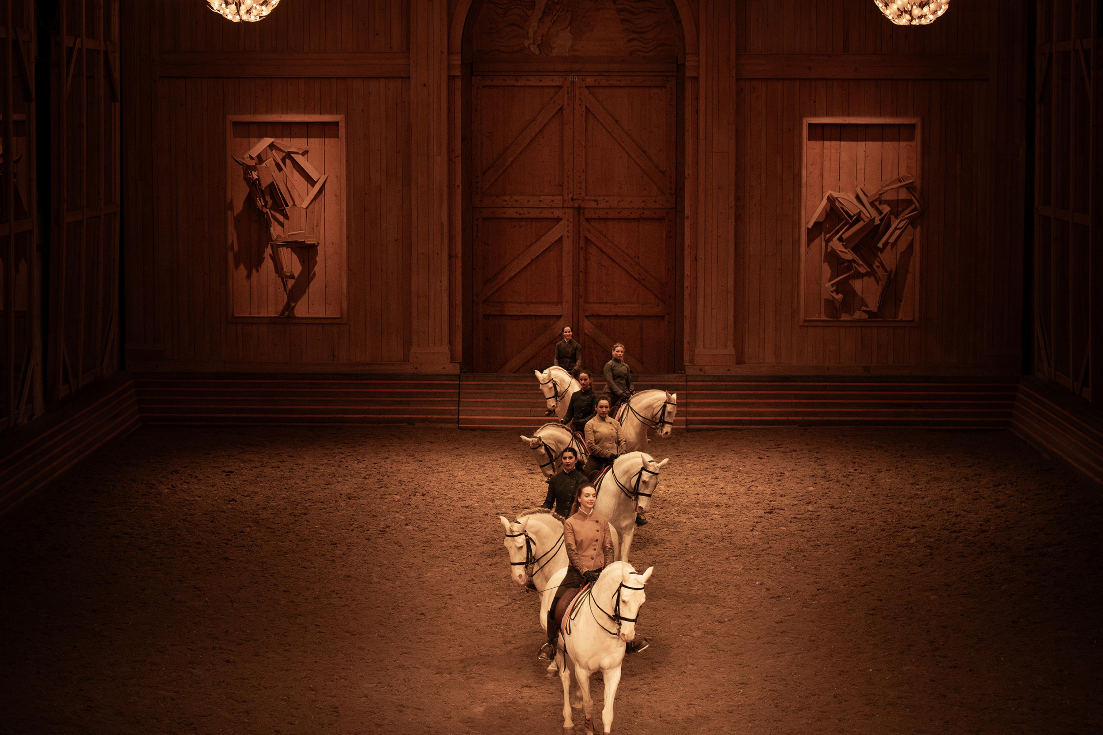 horses being ridden in a single file line inside an indoor arena with sand floor