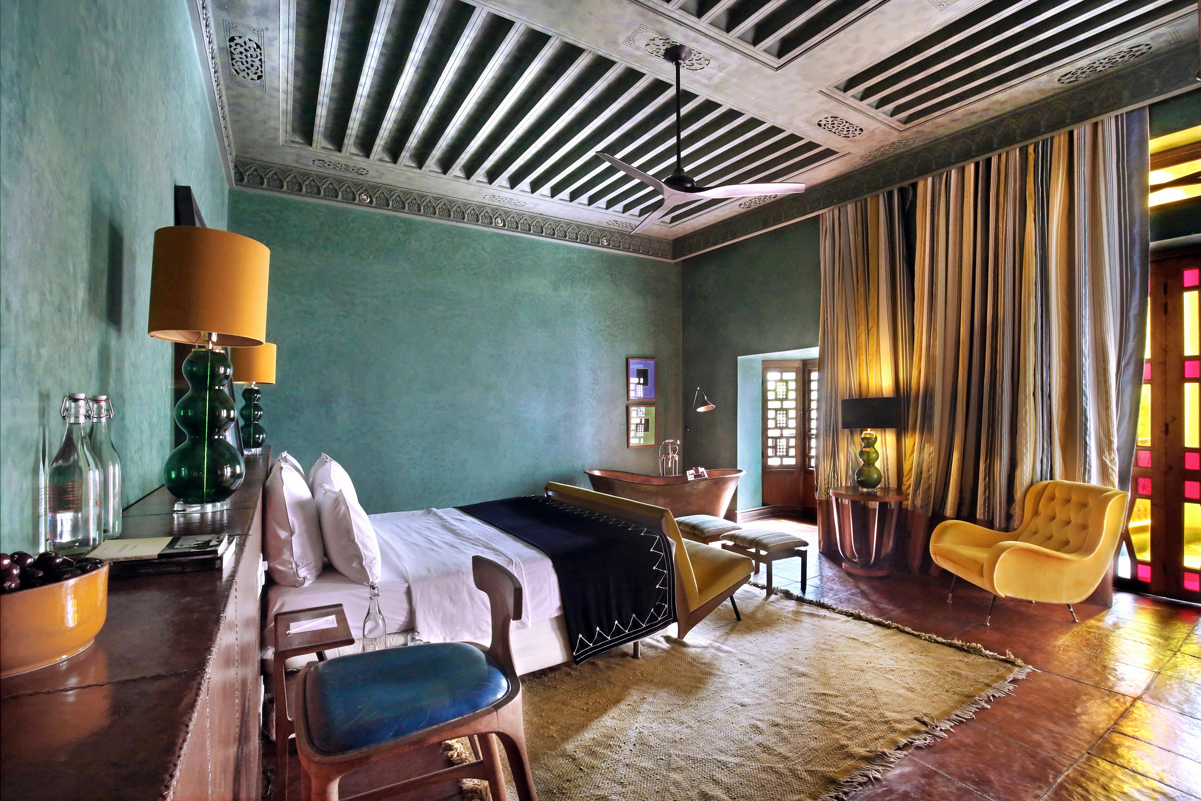 hotel room with wooden ceiling, green painted walls, terra cotta floor and a bed facing stained glass french doorway. there is a yellow chair and matching yellow lampshades on two lamps above bed