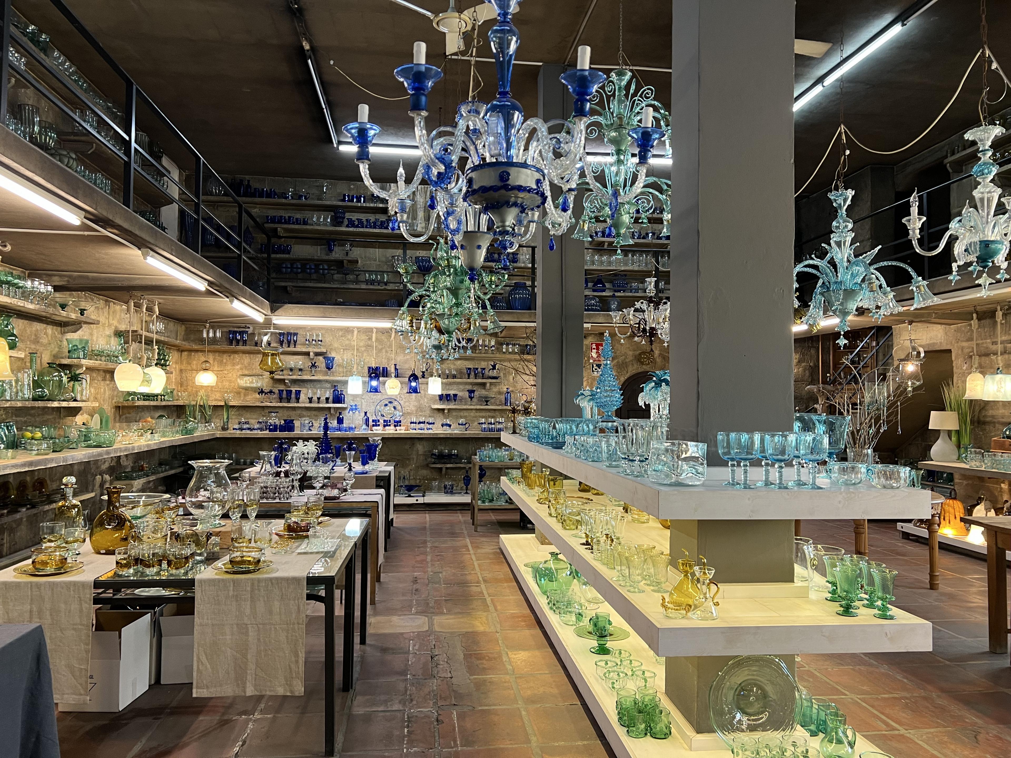 Store full of shelves and tables topped with glassware such as vases, wine glasses, platters, and glass chandeliers hanging above 