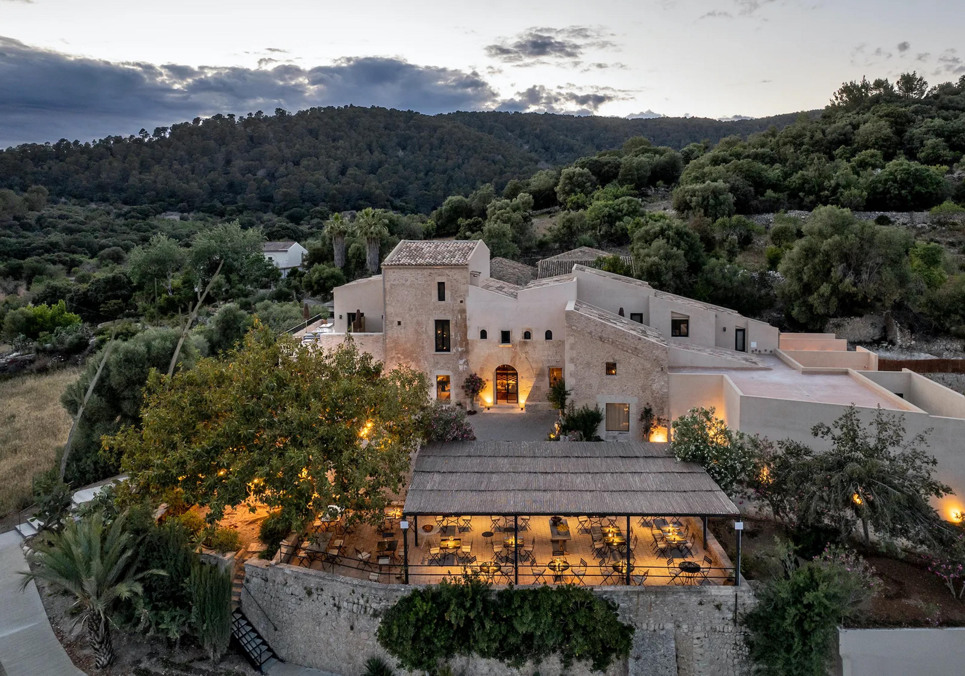 mediterranean-climate hill with multi-story stone / stucco hotel building seen from above at dusk with a terrace in front lit up for dinner