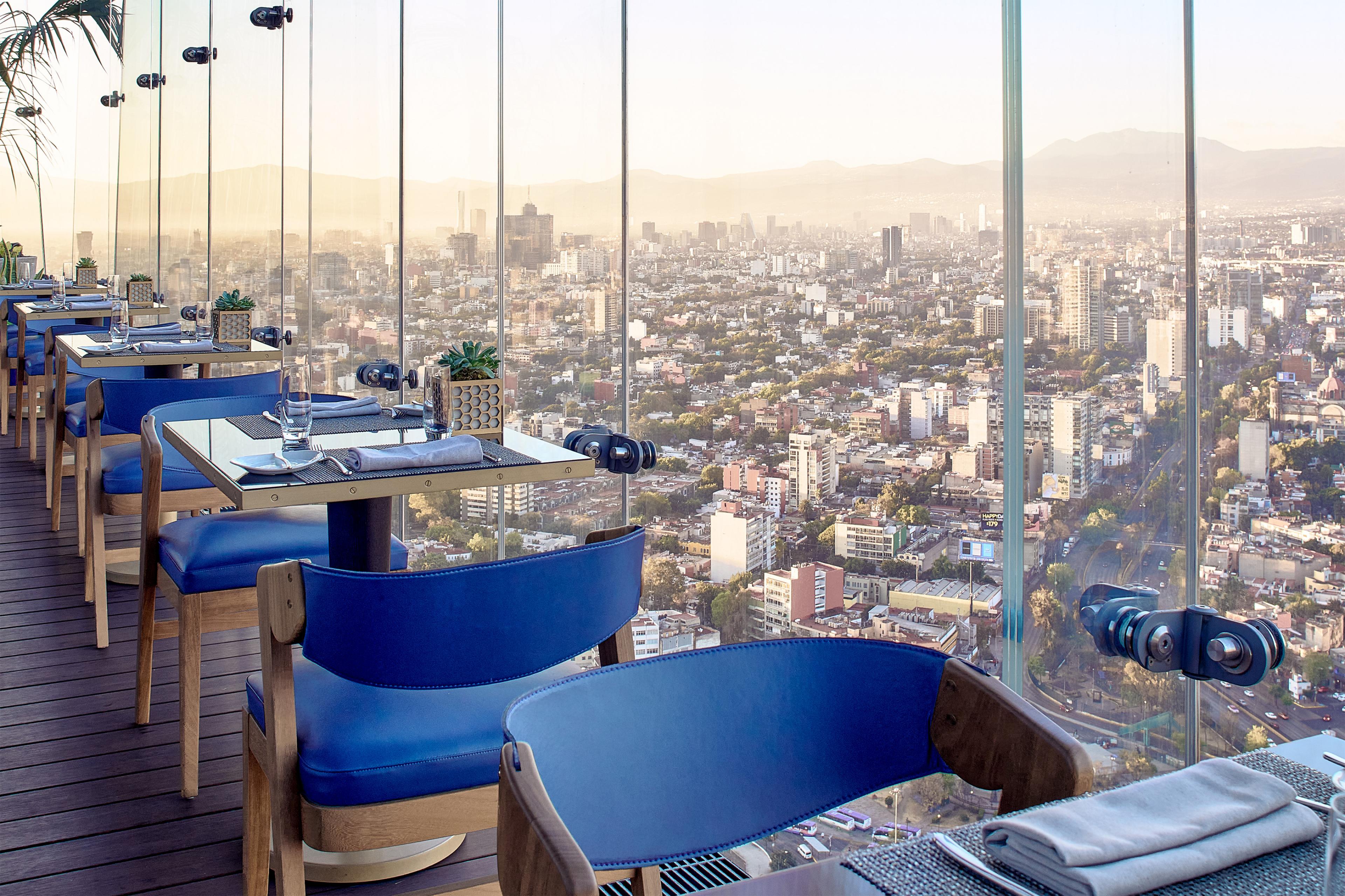 tables for two with blue chairs set along floor to ceiling windows overlooking a city