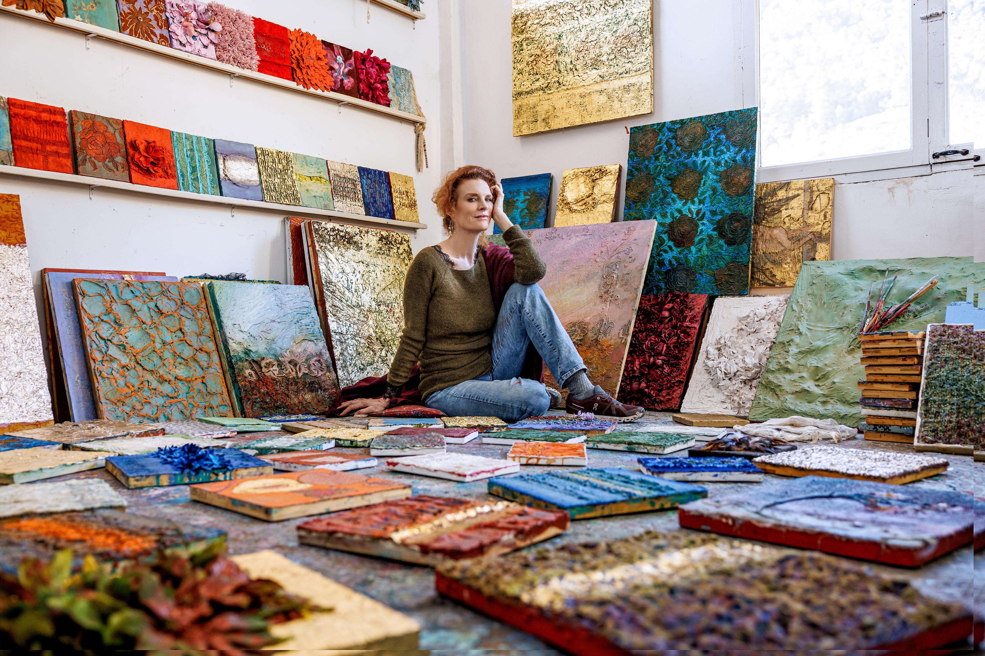 woman with red hair sitting on floor surrounded by colorful abstract paintings arranged on floor and stacked by wall