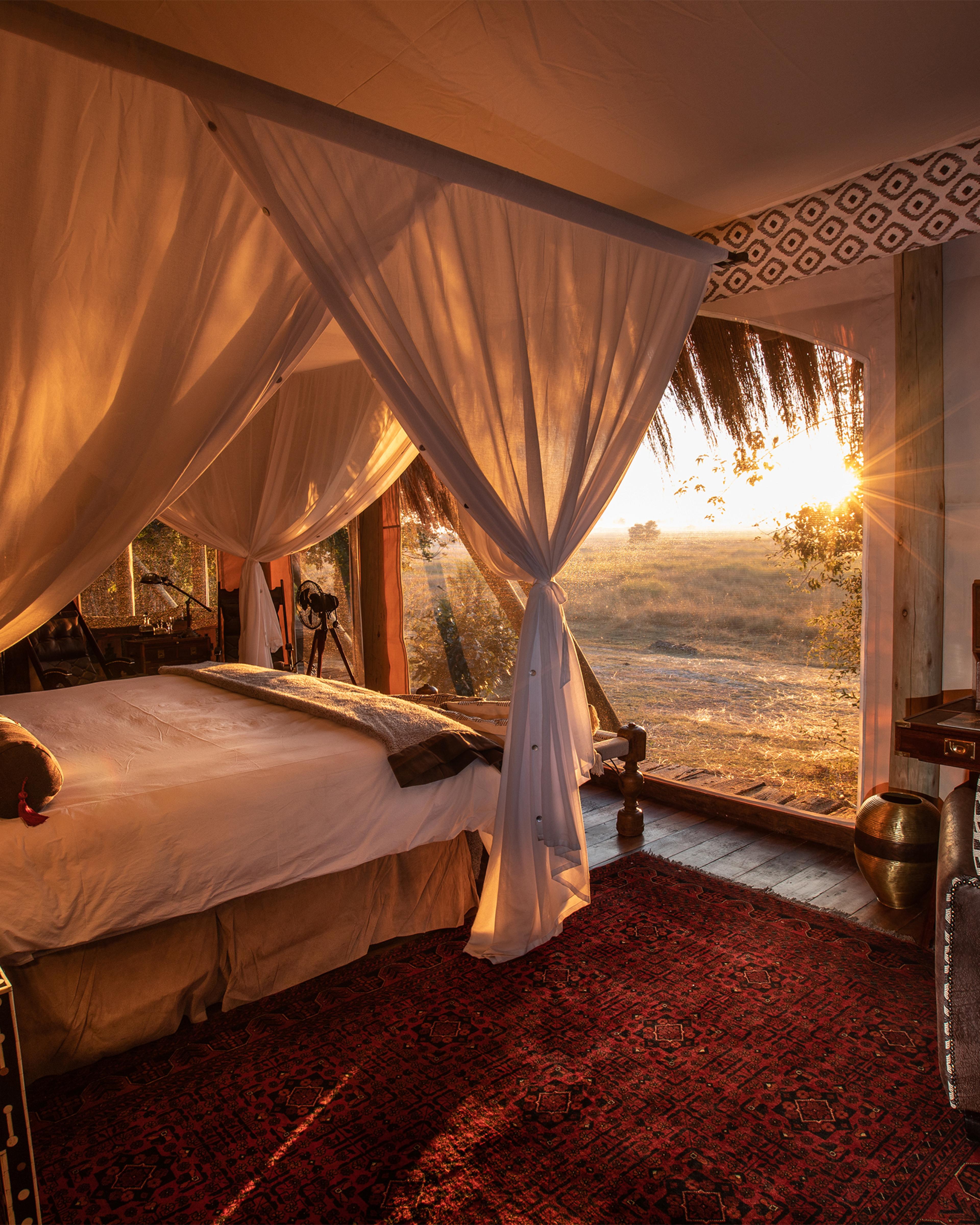 safari tent with bed on left and sunset viewed through window in canvas wall on right