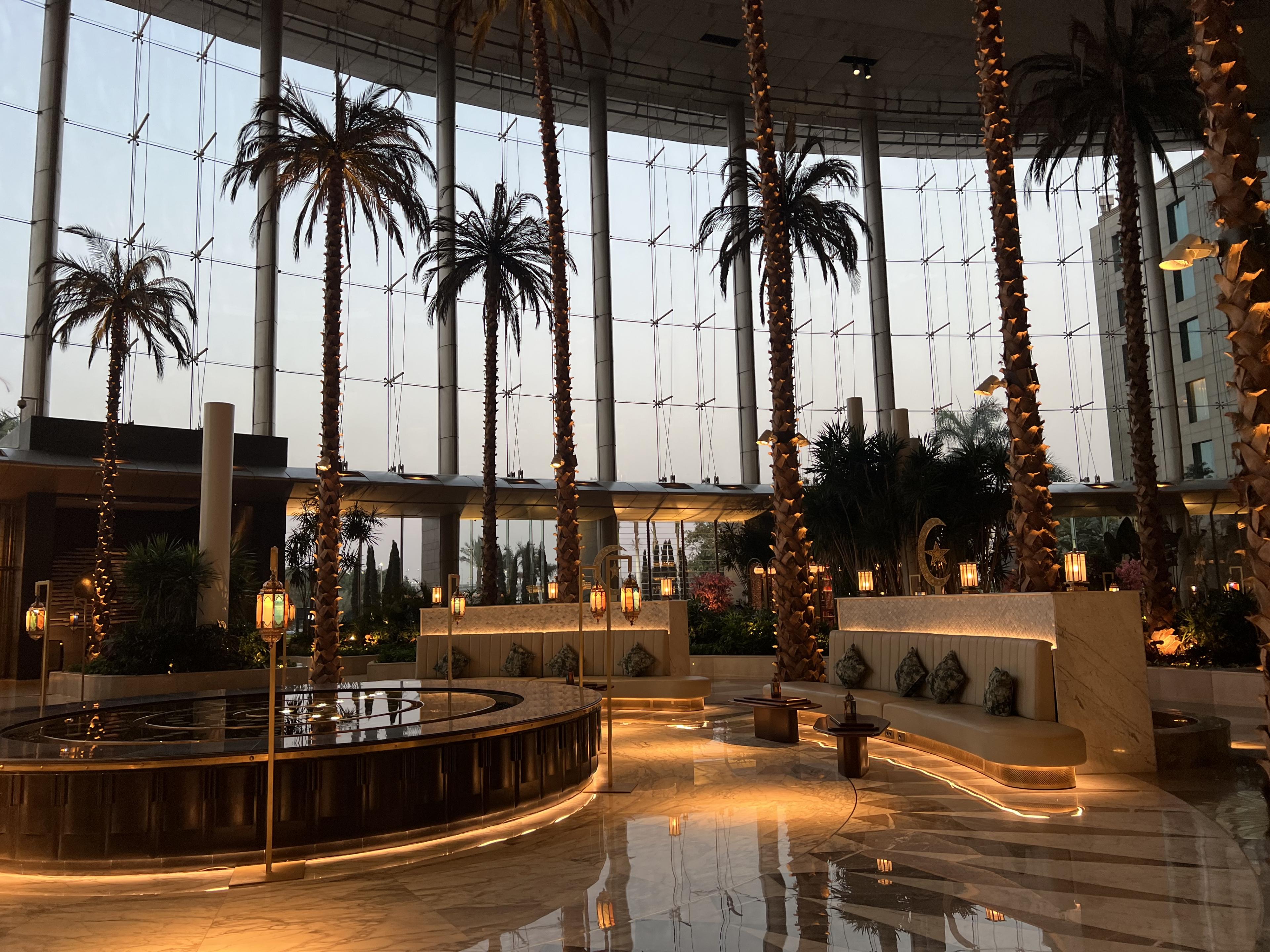 Hotel lobby with palm trees throughout in front of a glass wall with a view of outside 