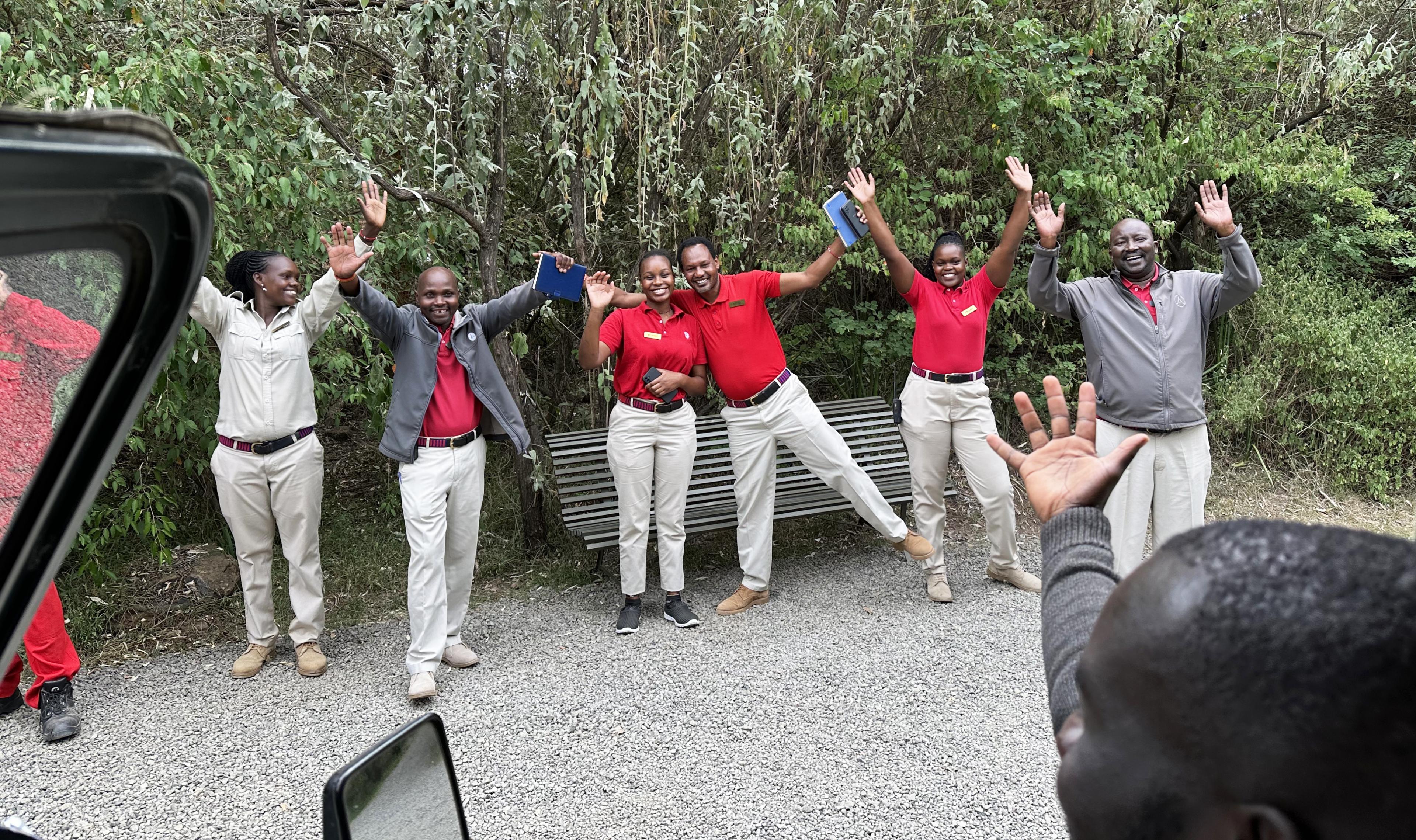 a team of safari lodge staff wearing red shirts and khaki pants waves goodbye 