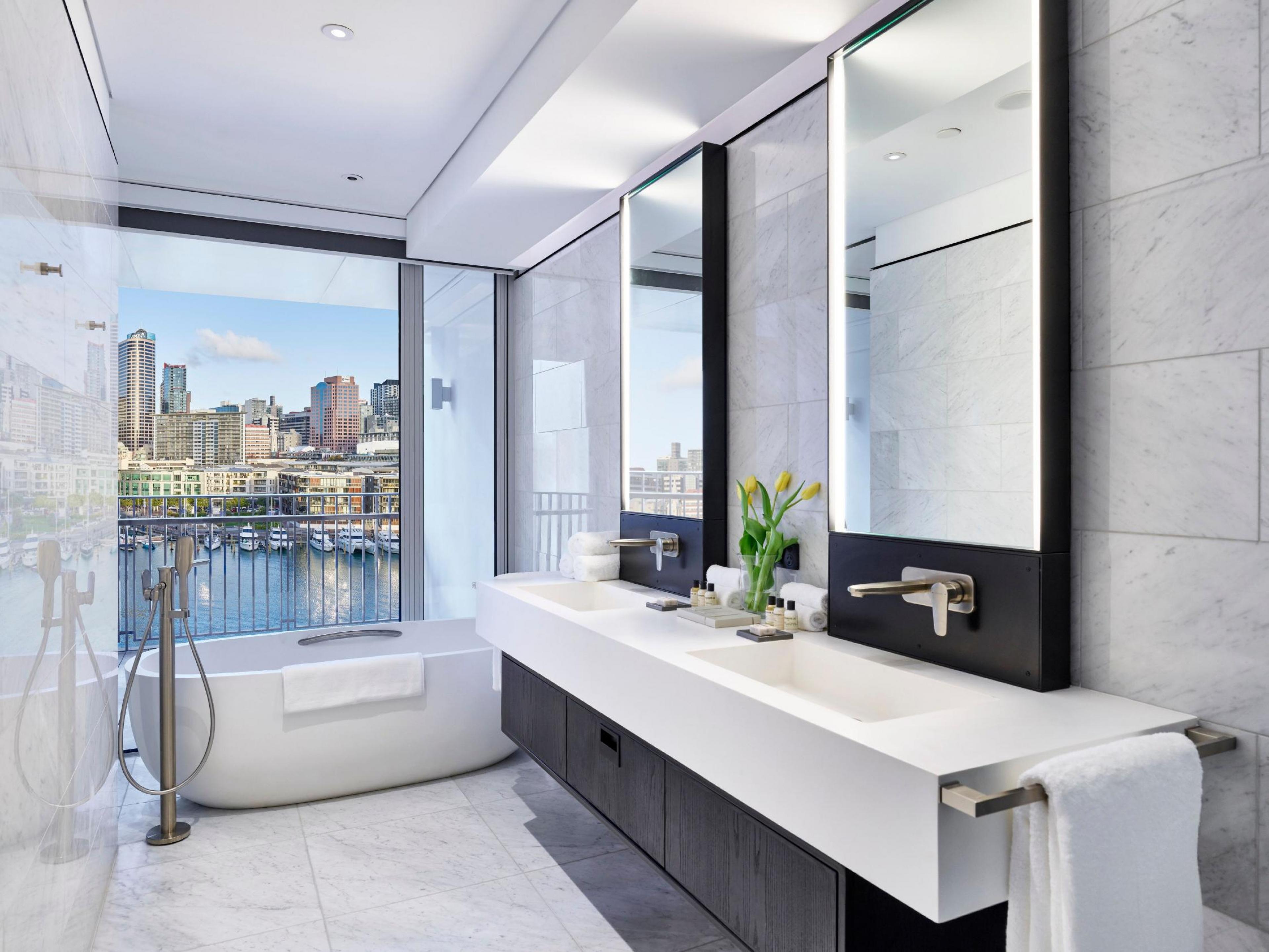 A bathroom with gray and white tiled flooring and wall with a white sink and two mirrors along the wall and a large soaking tub in the back in front of a window overlooking Auckland 