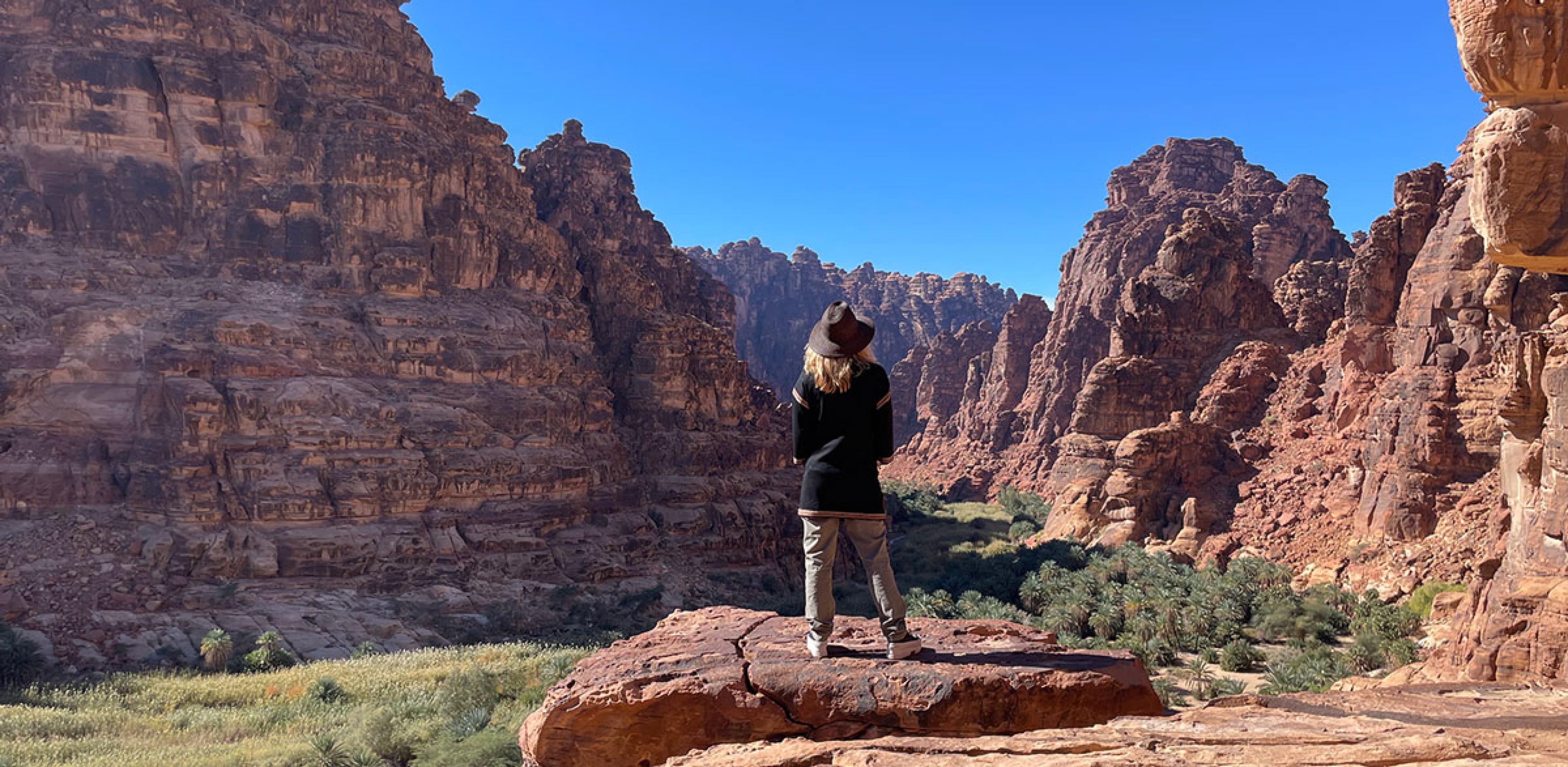 woman stands on edge of rocky cliff looking out over red rock canyon in saudi arabia