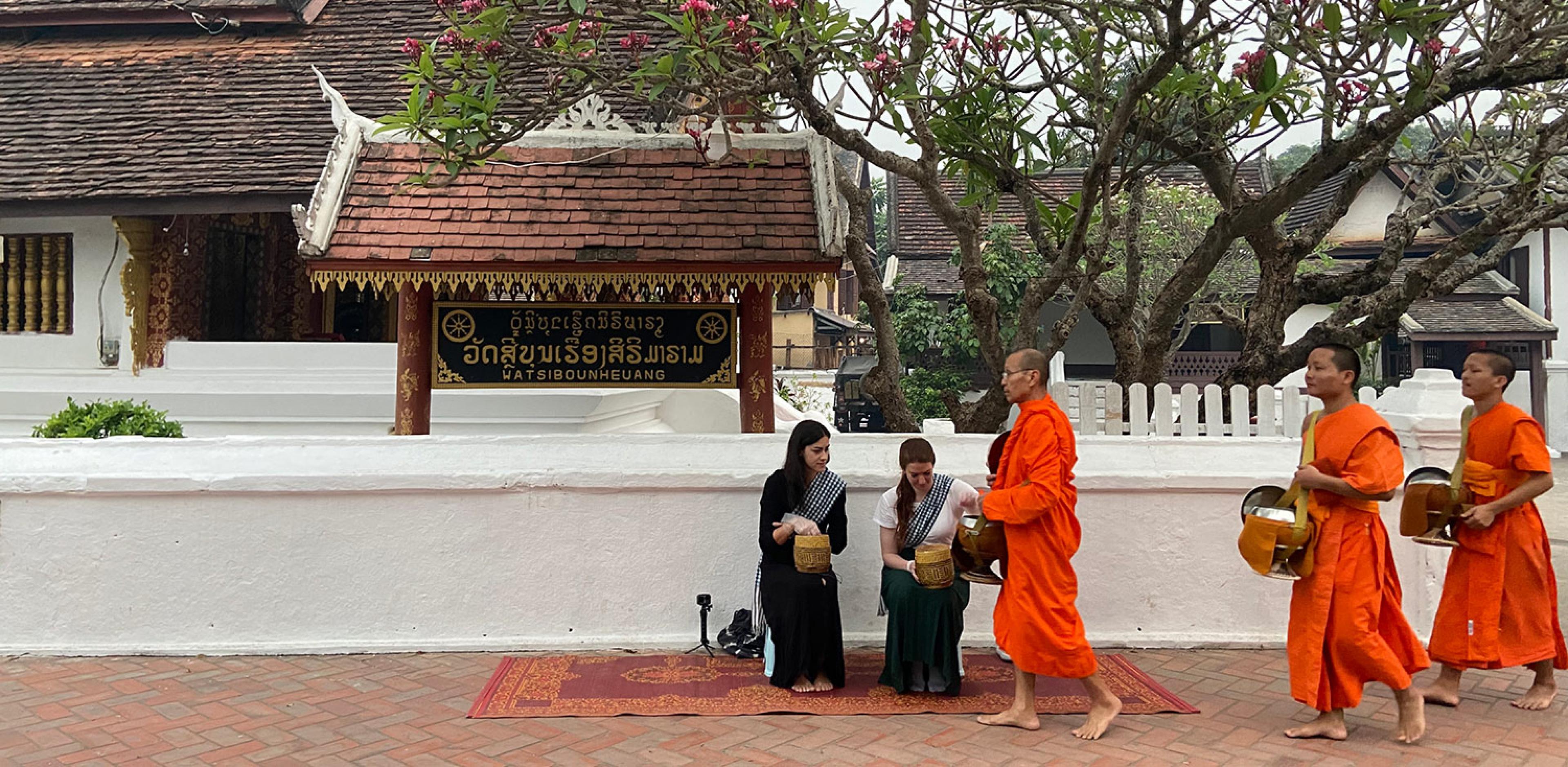 two women kneeling on street in luang prabang as monks wearing orange robes walk by 