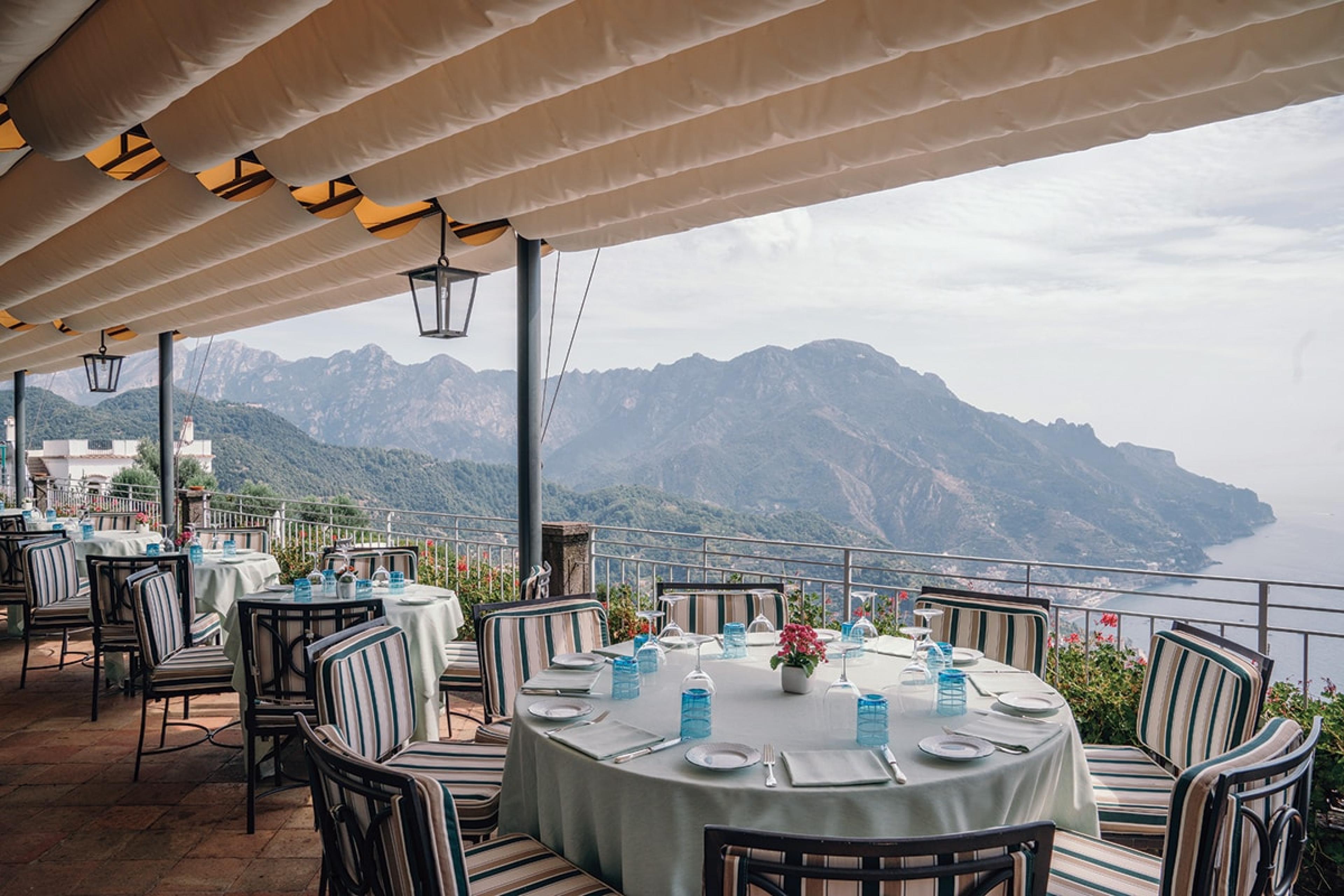 terrace with table with white tablecloth and blue water glasses overlooking amalfi coast with its hills leading down to the sea