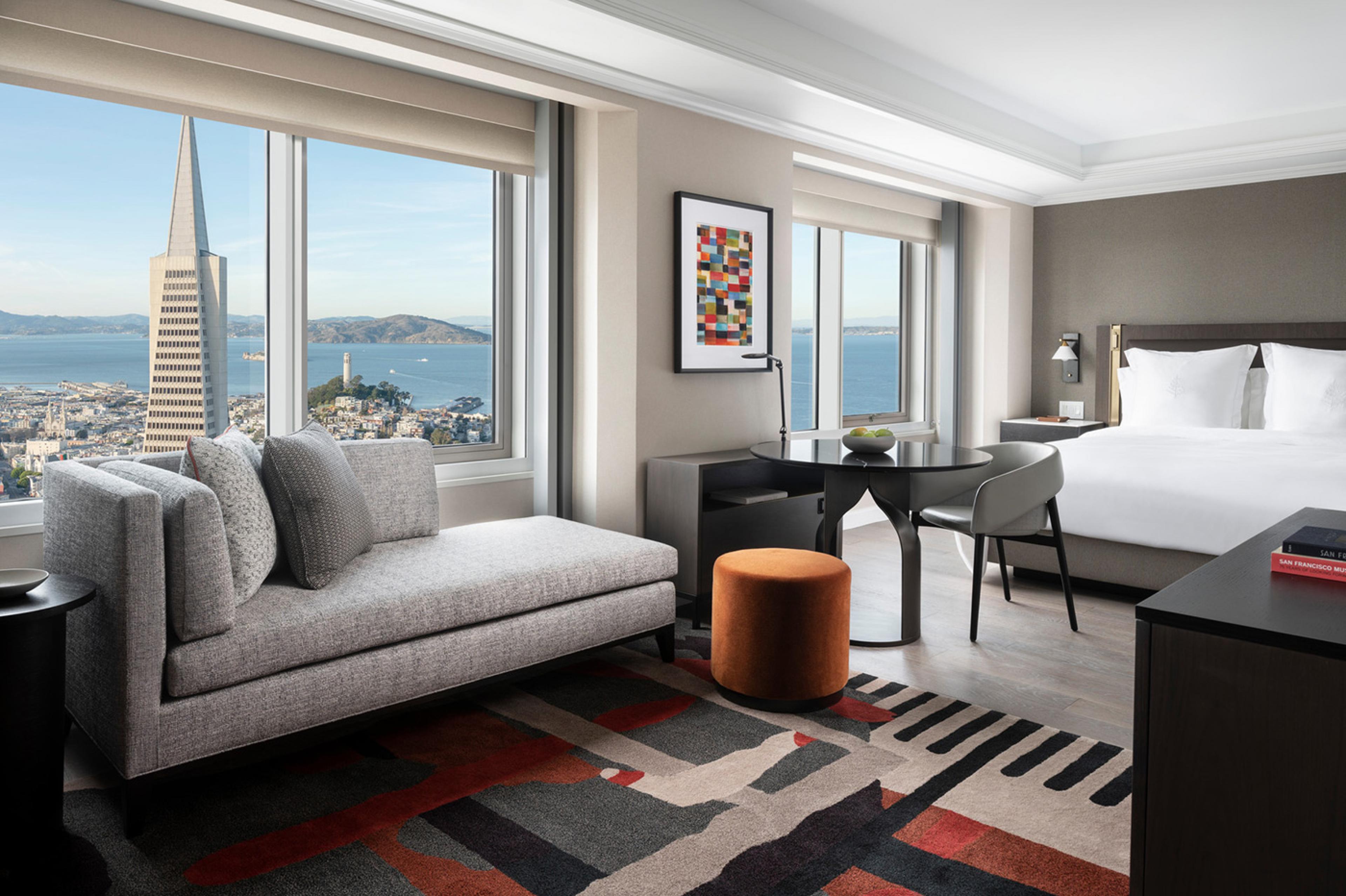 hotel room with gray walls and gray and white furniture with red accent color, with big window looking out to san francisco skyline with transamerica pyramid